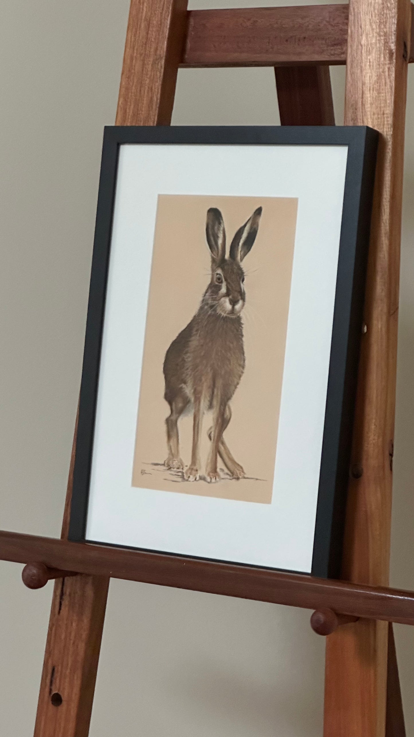 Framed illustration of a rabbit on a wooden easel against a plain wall.