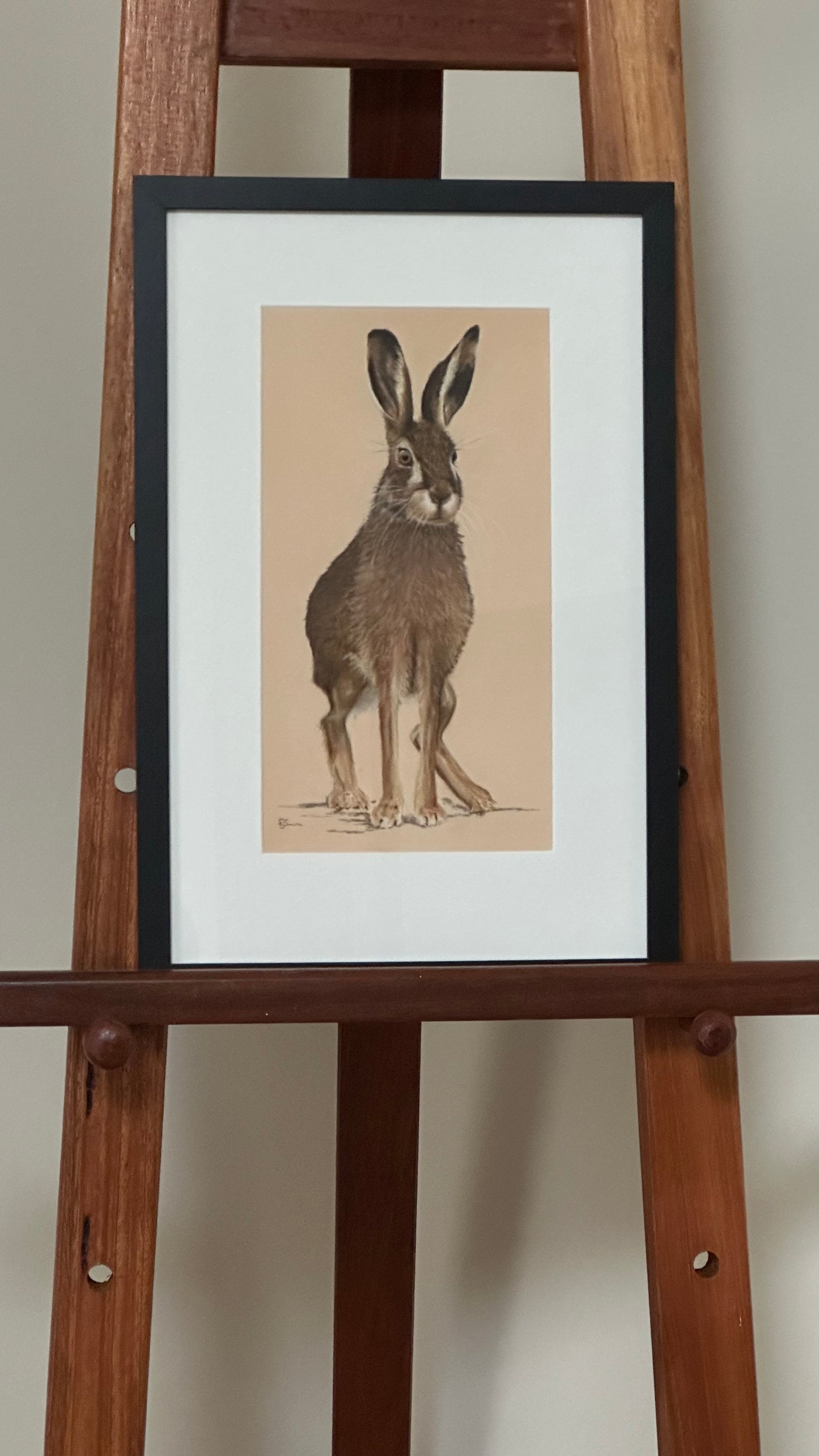 Framed illustration of a rabbit on a wooden easel against a plain wall.
