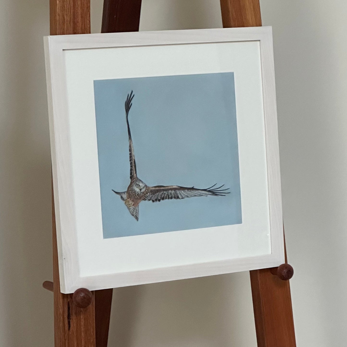 Framed artwork of a bird on a wooden easel against a plain wall.