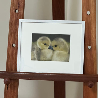Framed picture of a duckling on a wooden easel against a plain wall.