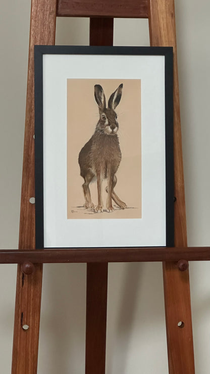Framed illustration of a rabbit on a wooden easel against a plain wall.