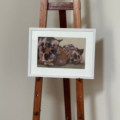 Framed picture of two pigs on a wooden easel against a plain wall.