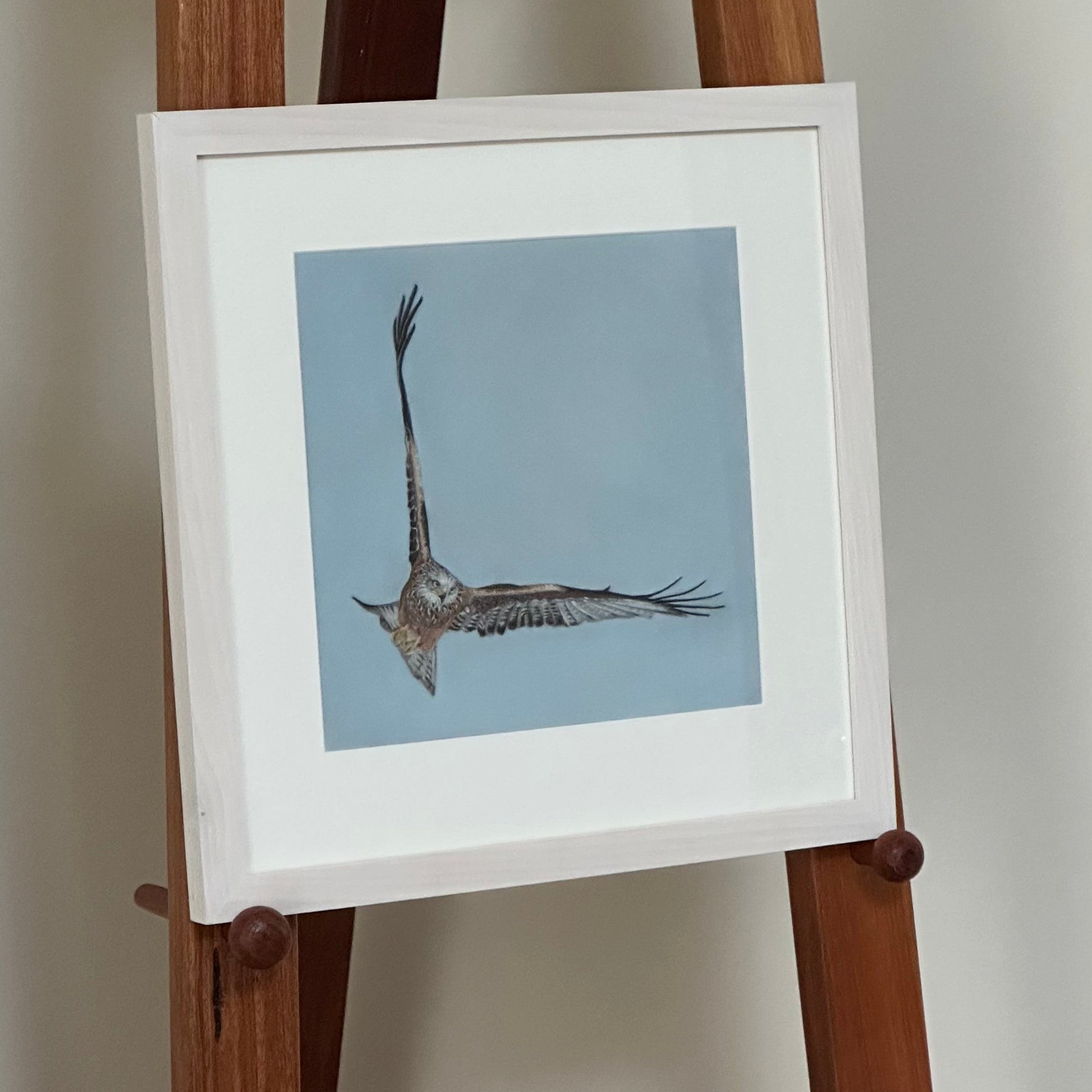 Framed artwork of a bird on a wooden easel against a plain wall.