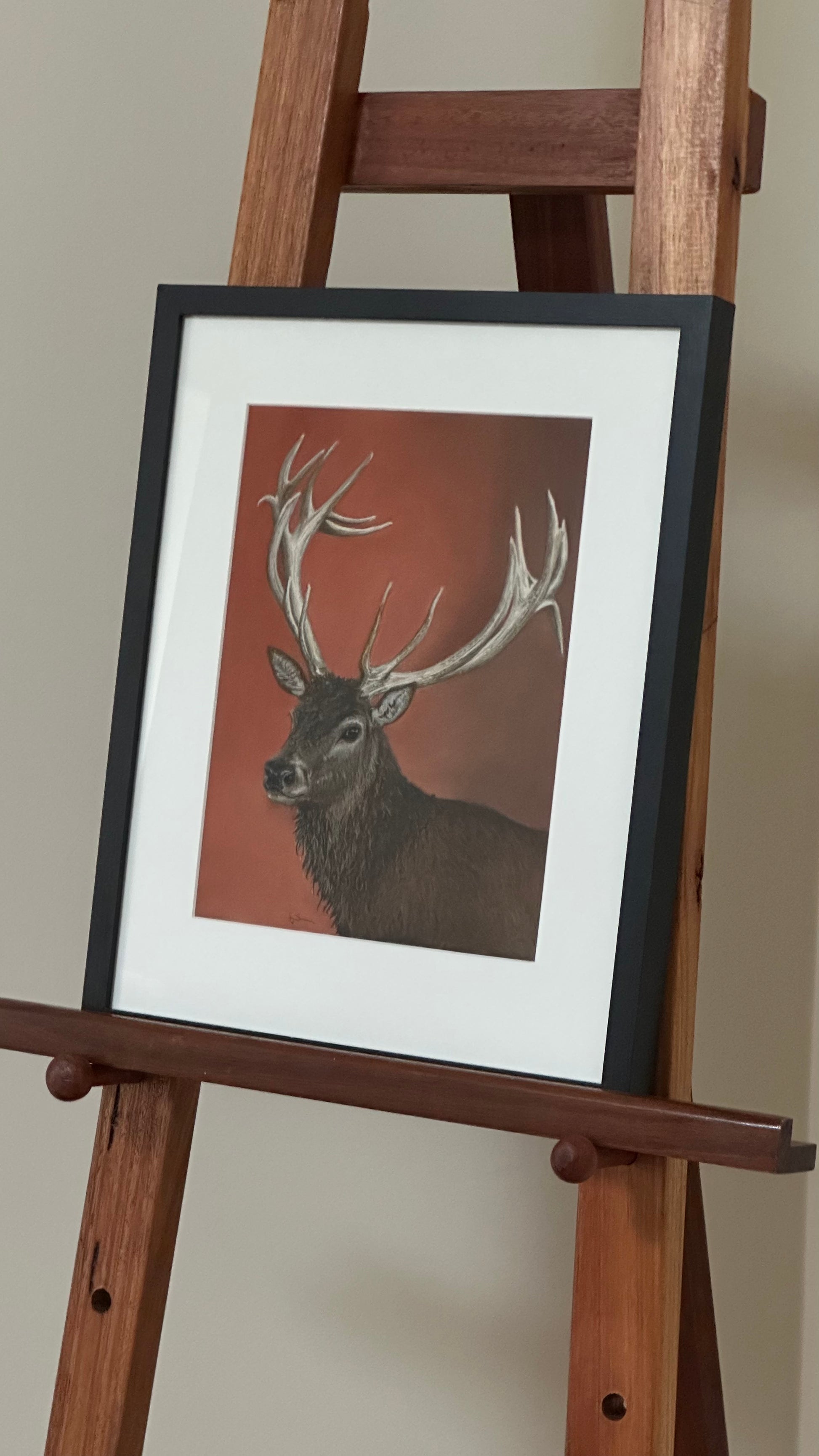 Framed artwork of a deer on a wooden easel against a plain wall.