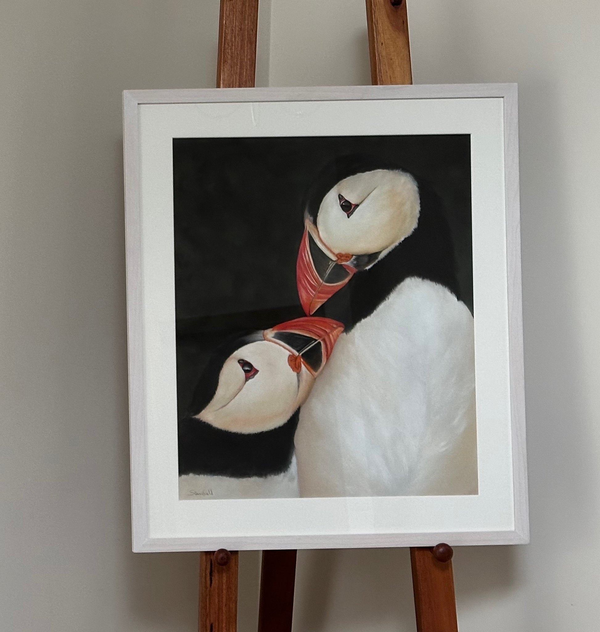 Framed artwork of two puffins on a black background, displayed on an easel.