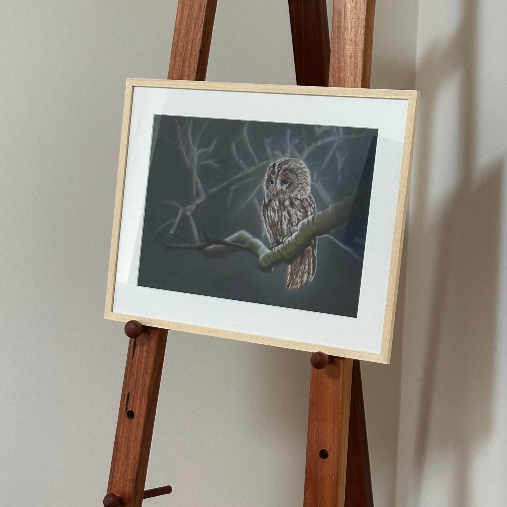Framed artwork of an owl on a wooden easel against a plain wall.