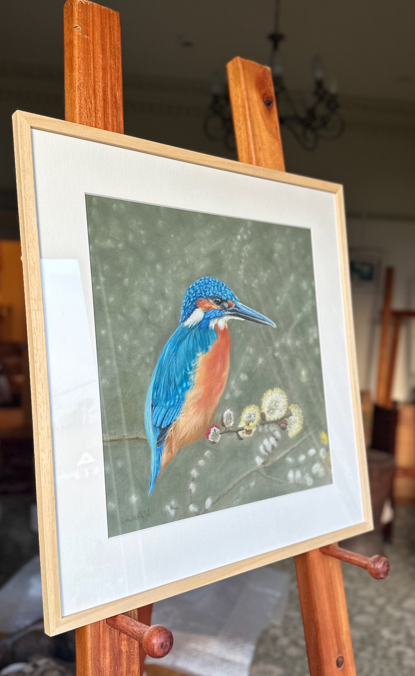 Framed artwork of a blue and orange kingfisher on an easel in a room 