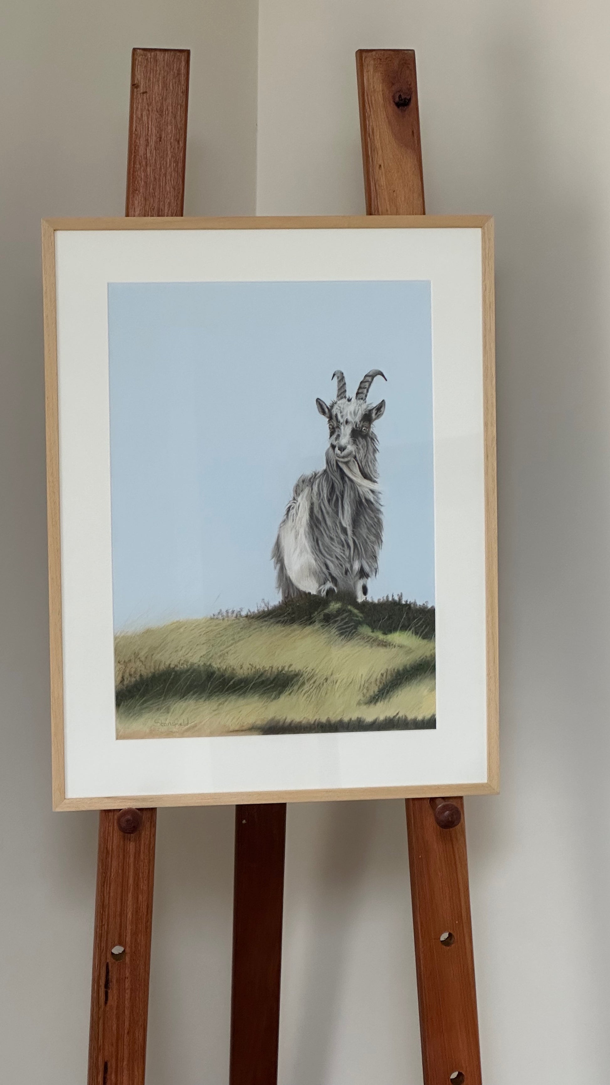 Framed artwork of a goat on a grassy hill with a wooden easel against a plain wall.