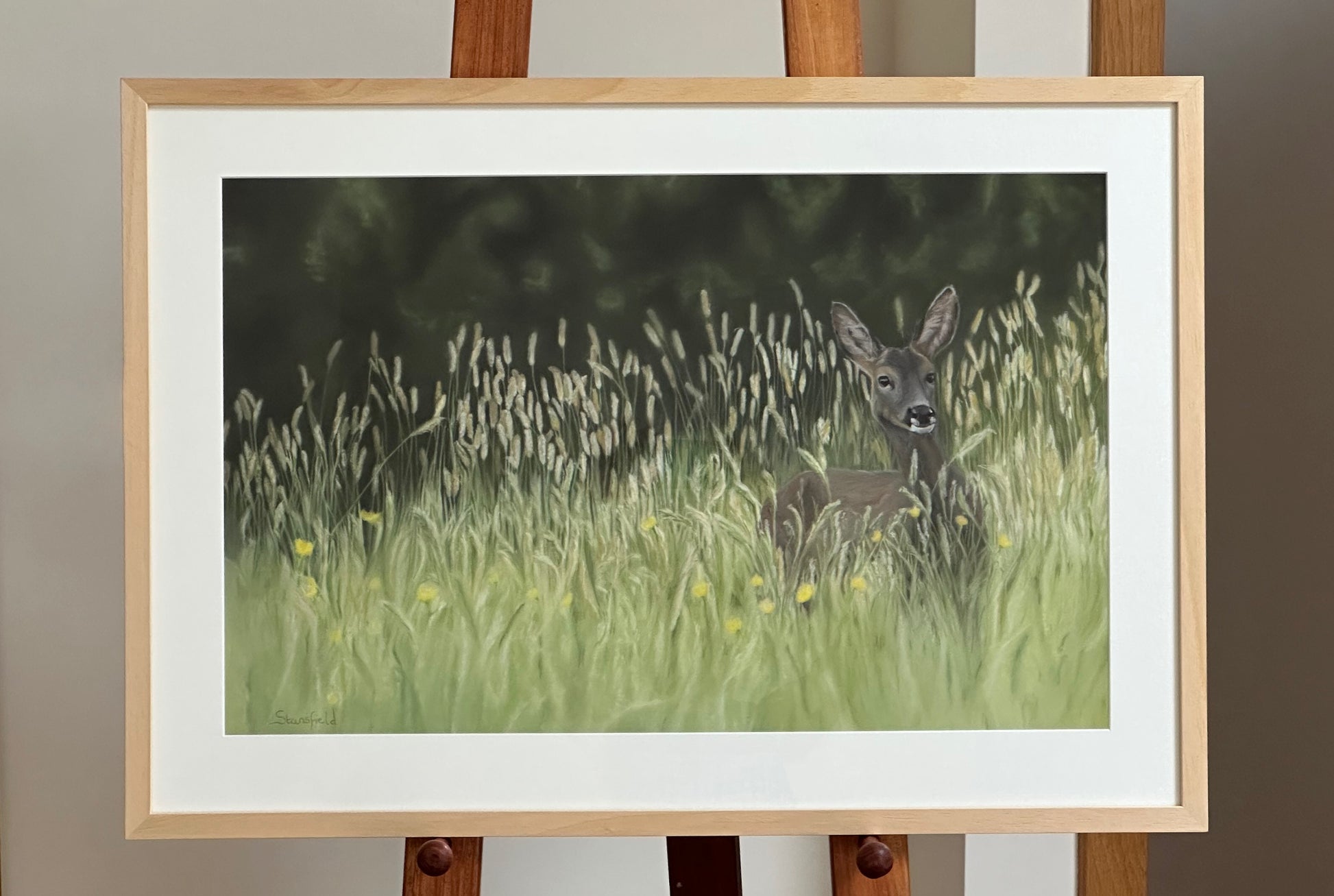 Framed artwork of a deer in a grassy field on an easel.