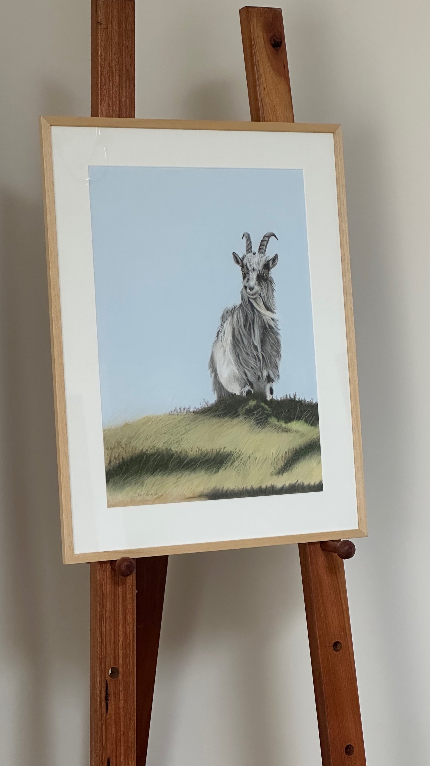 Framed artwork of a goat on a grassy hill displayed on a wooden easel.