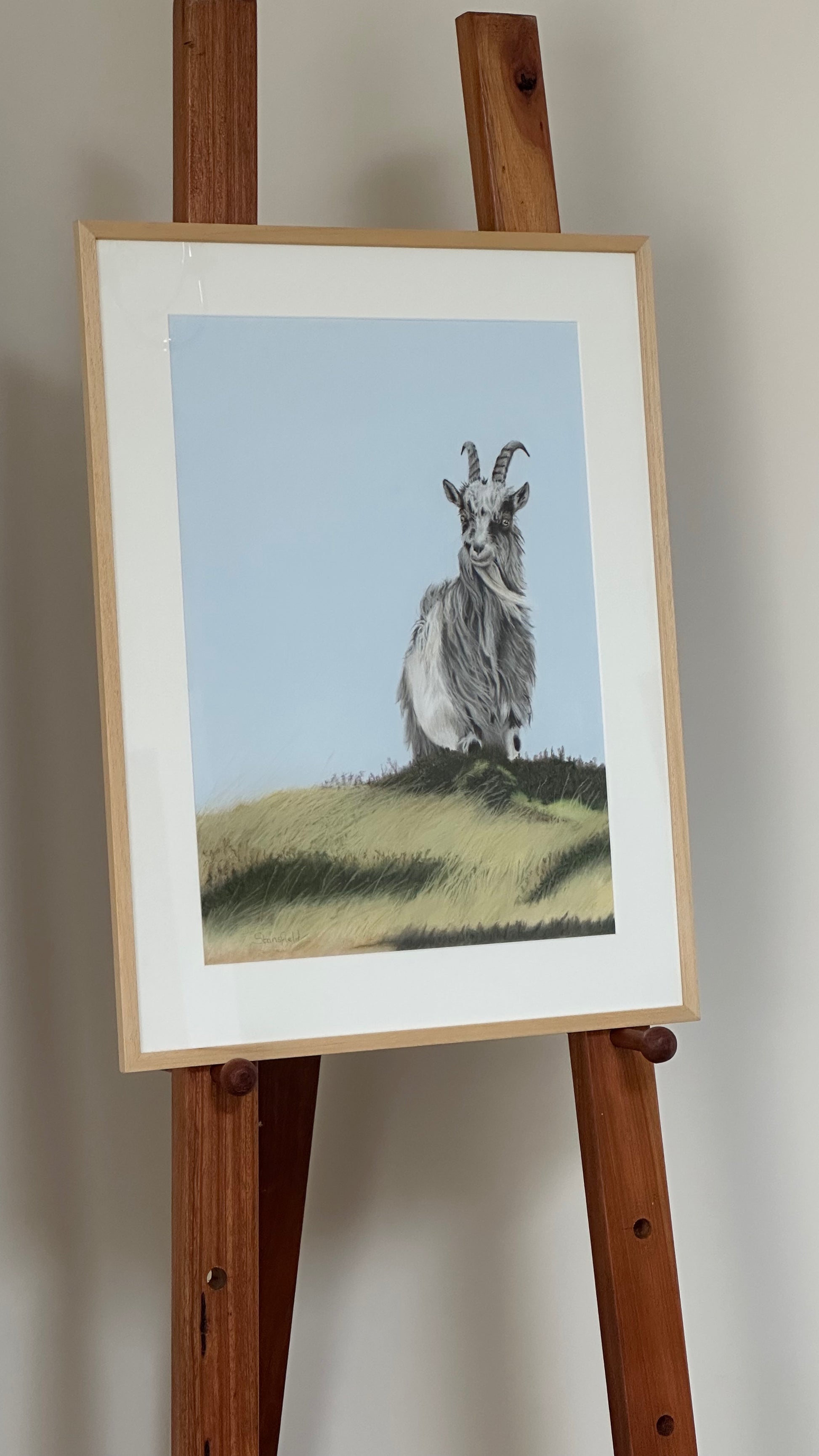 Framed artwork of a goat on a grassy hill displayed on a wooden easel.